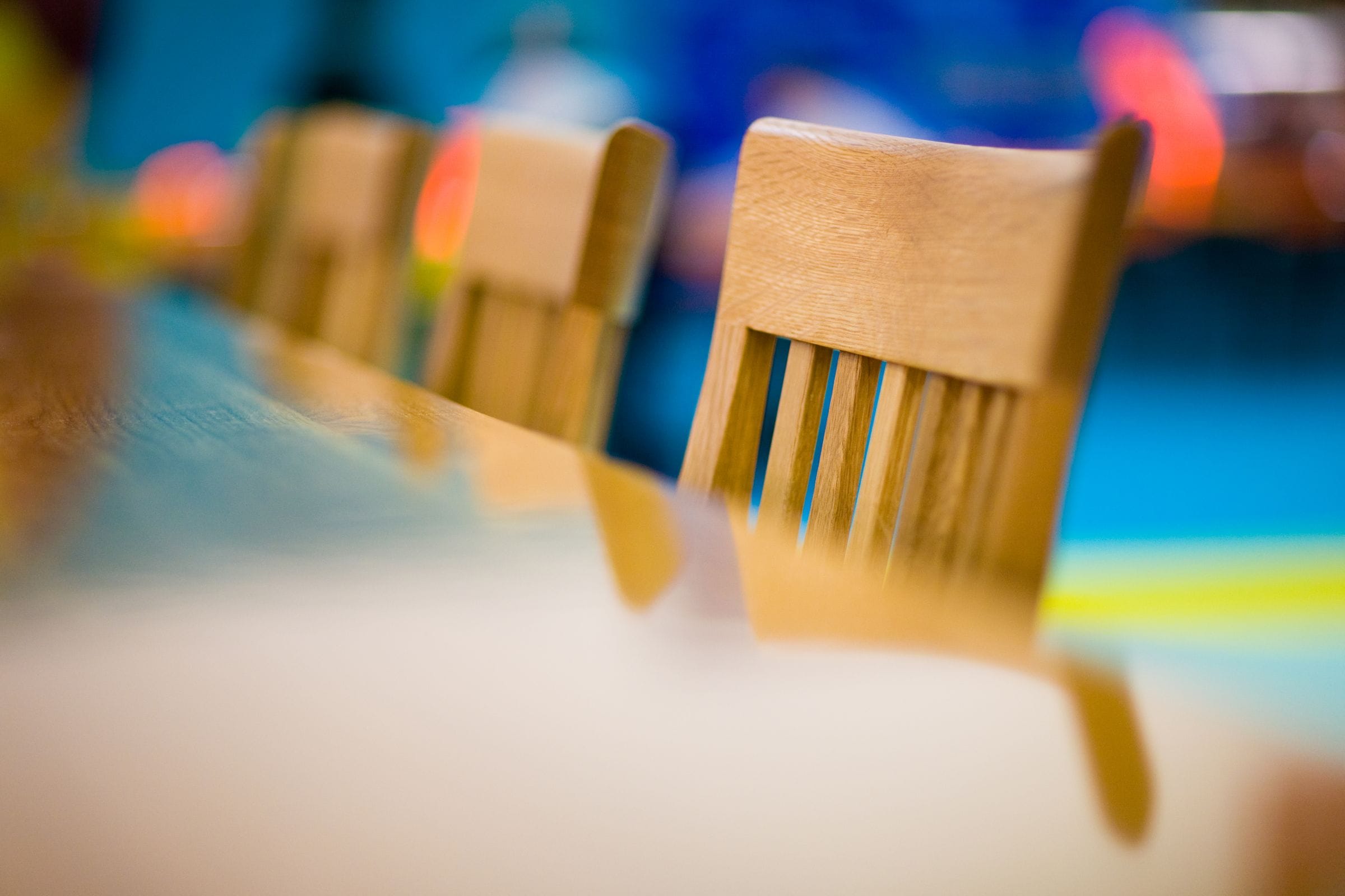 Oak slat-back dining chairs along a table against a colourful blurred background
