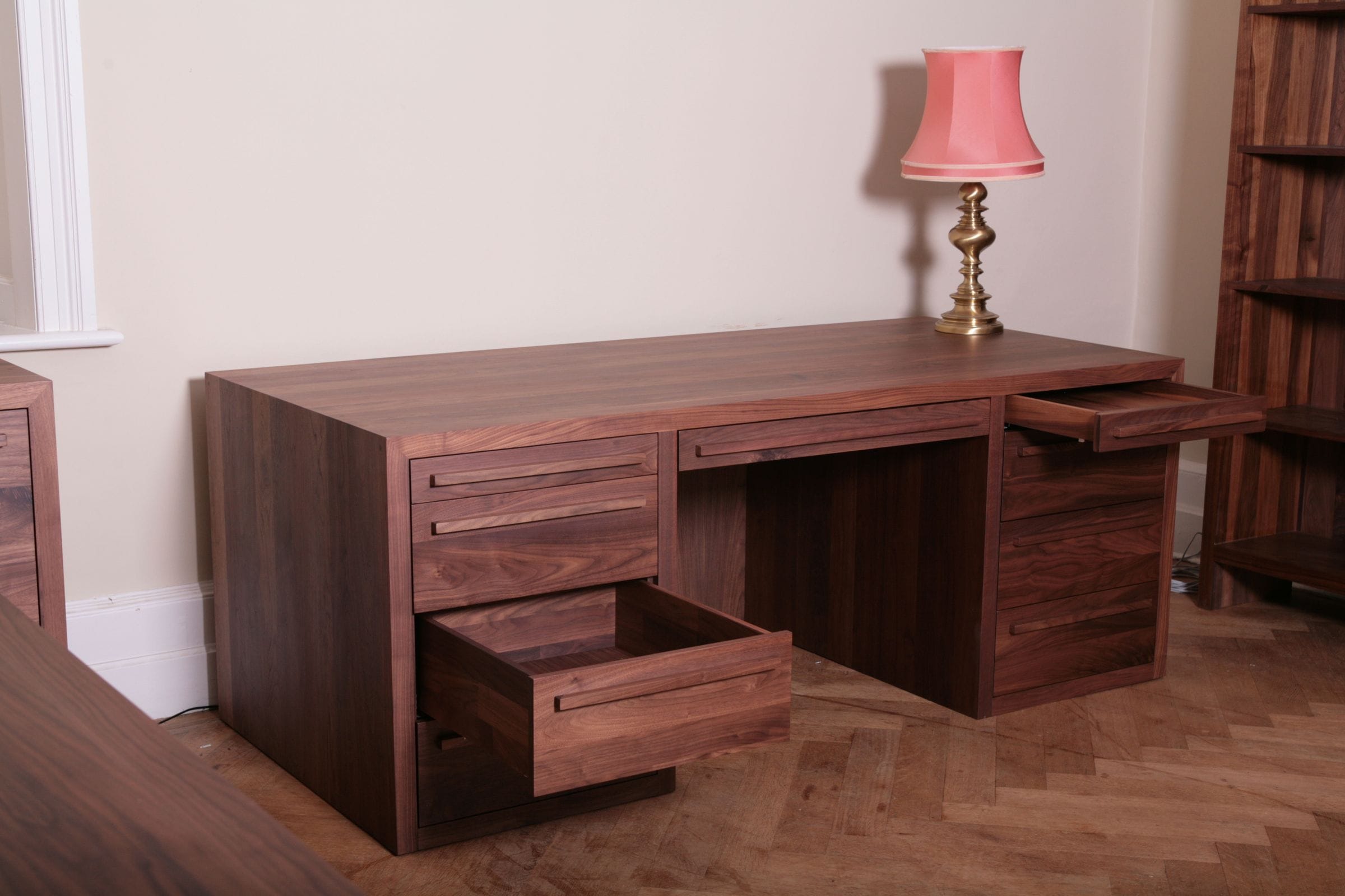 Walnut pedestal desk with drawers open, beside a pink lamp on parquet flooring