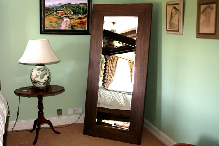 Full-length walnut-framed mirror leaning against a bedroom wall beside a table lamp
