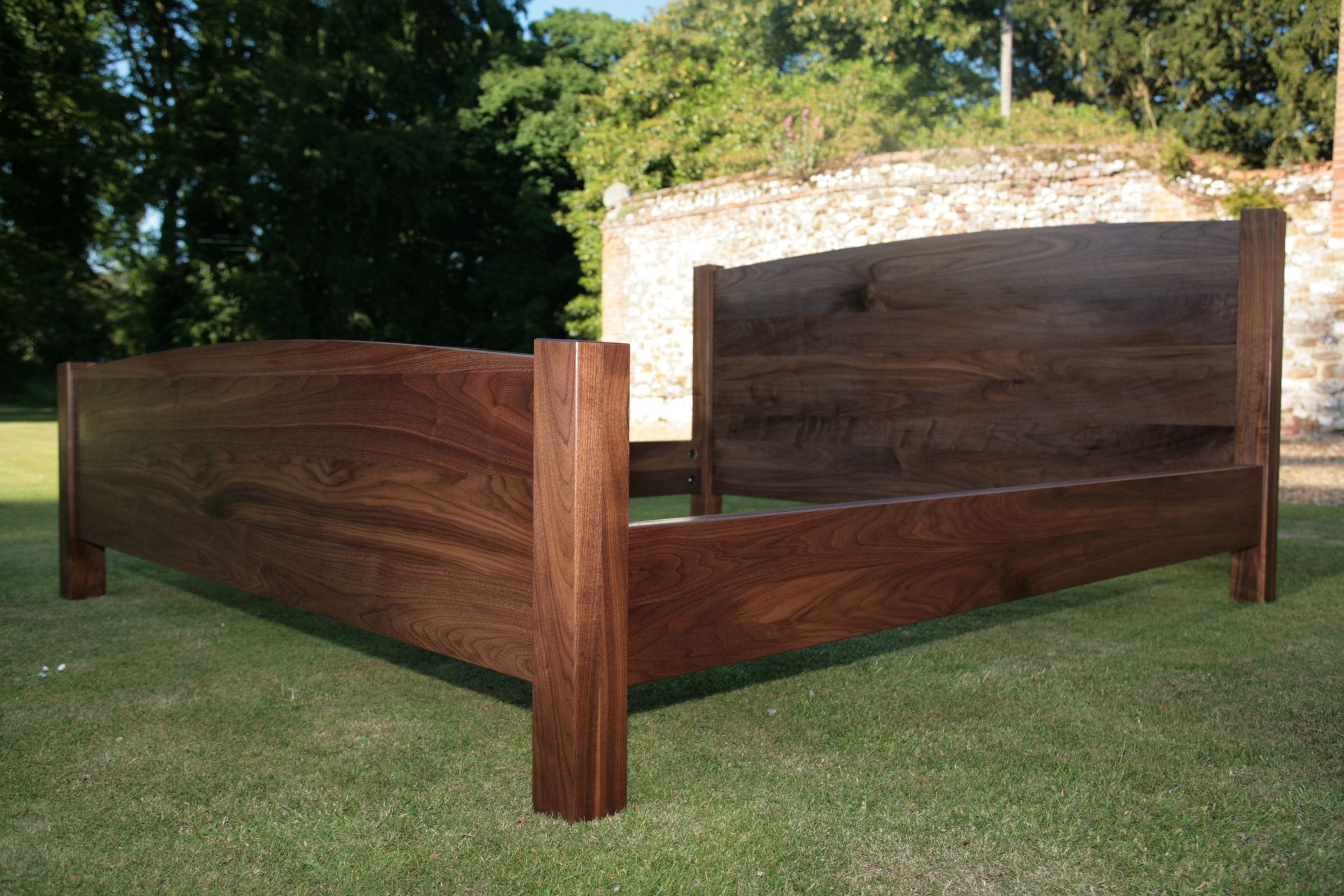 Walnut bed frame with curved headboard photographed on a lawn by a flint wall