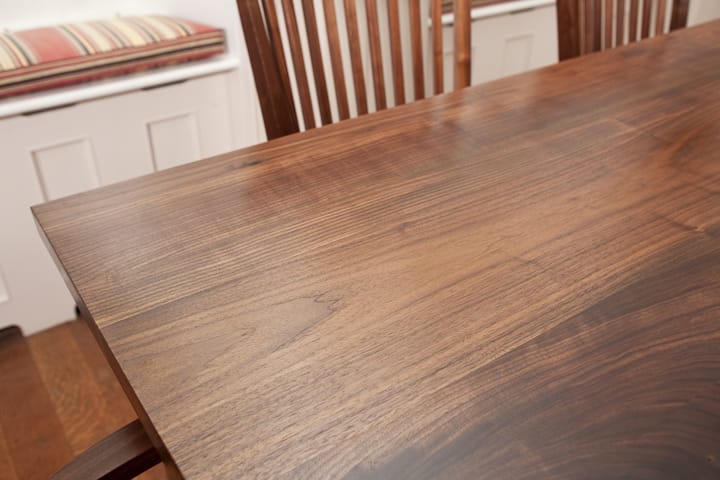 Close-up of a walnut dining tabletop showing rich grain, with slatted chairs behind