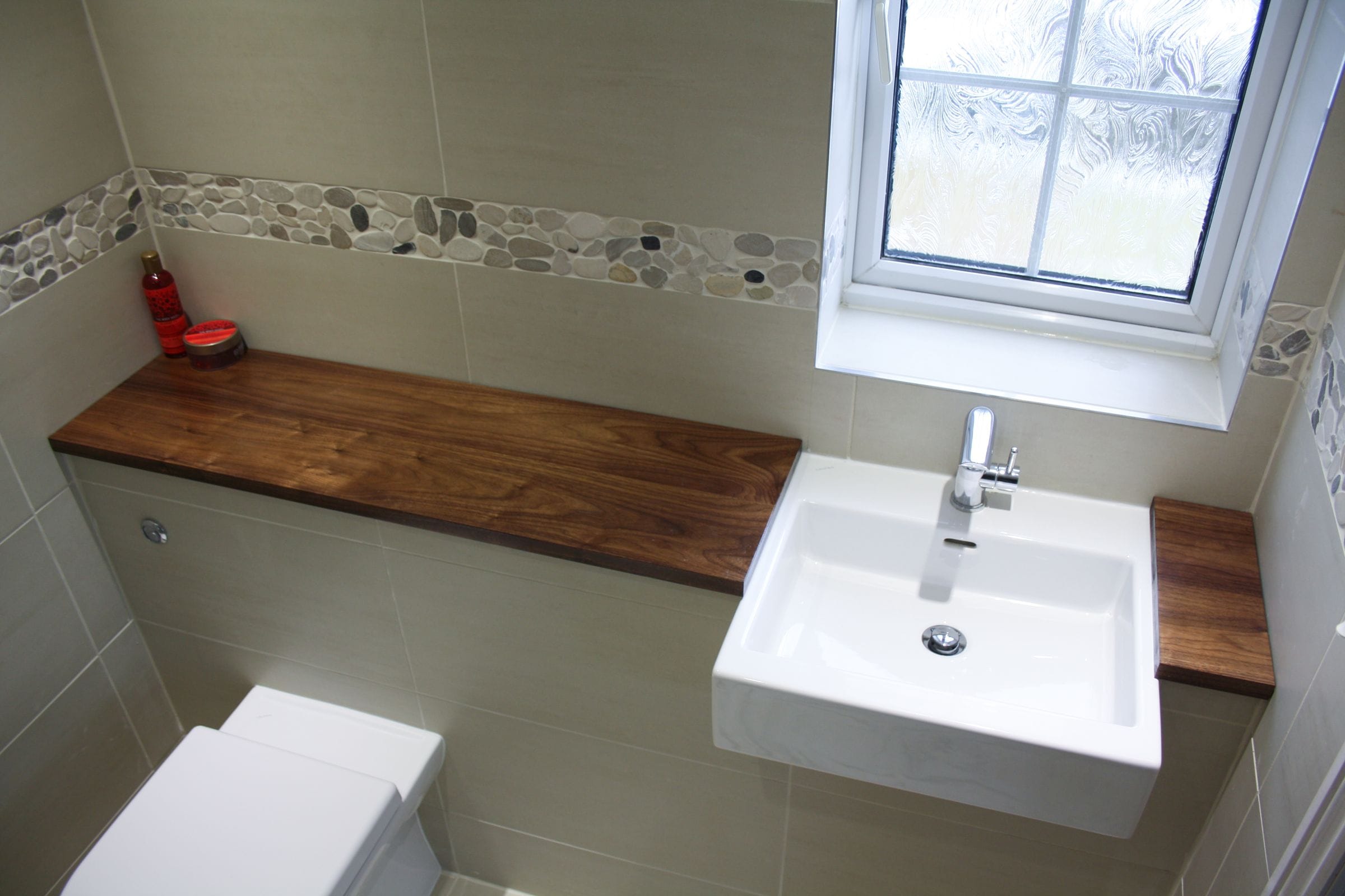 Walnut shelf and vanity tops around a wall-hung basin in a beige tiled bathroom