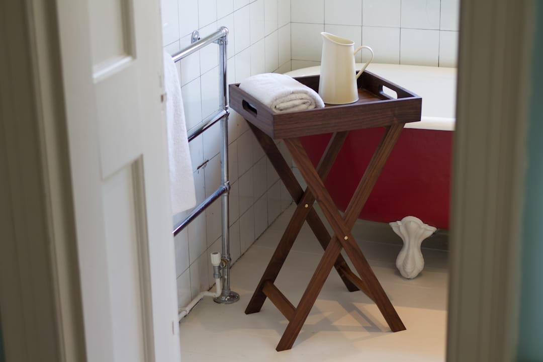 Walnut butler tray on a folding stand beside a roll-top bath, holding a jug and towel