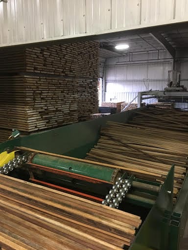 Sawn hardwood strips on a sawmill conveyor with tall stacks of boards behind
