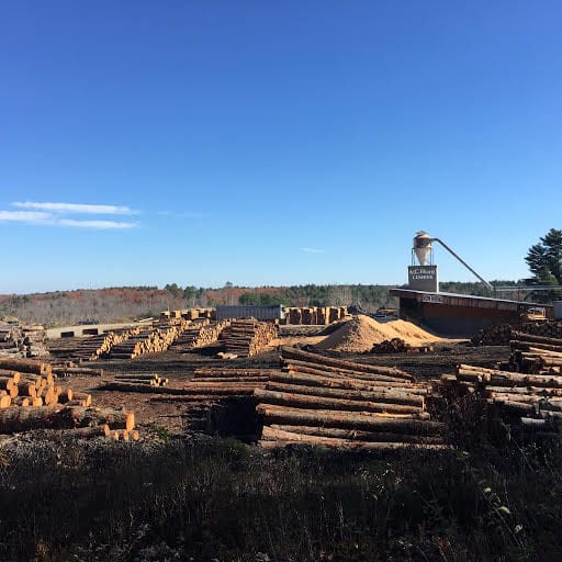Rows of stacked logs and a sawdust pile at a New England timber yard