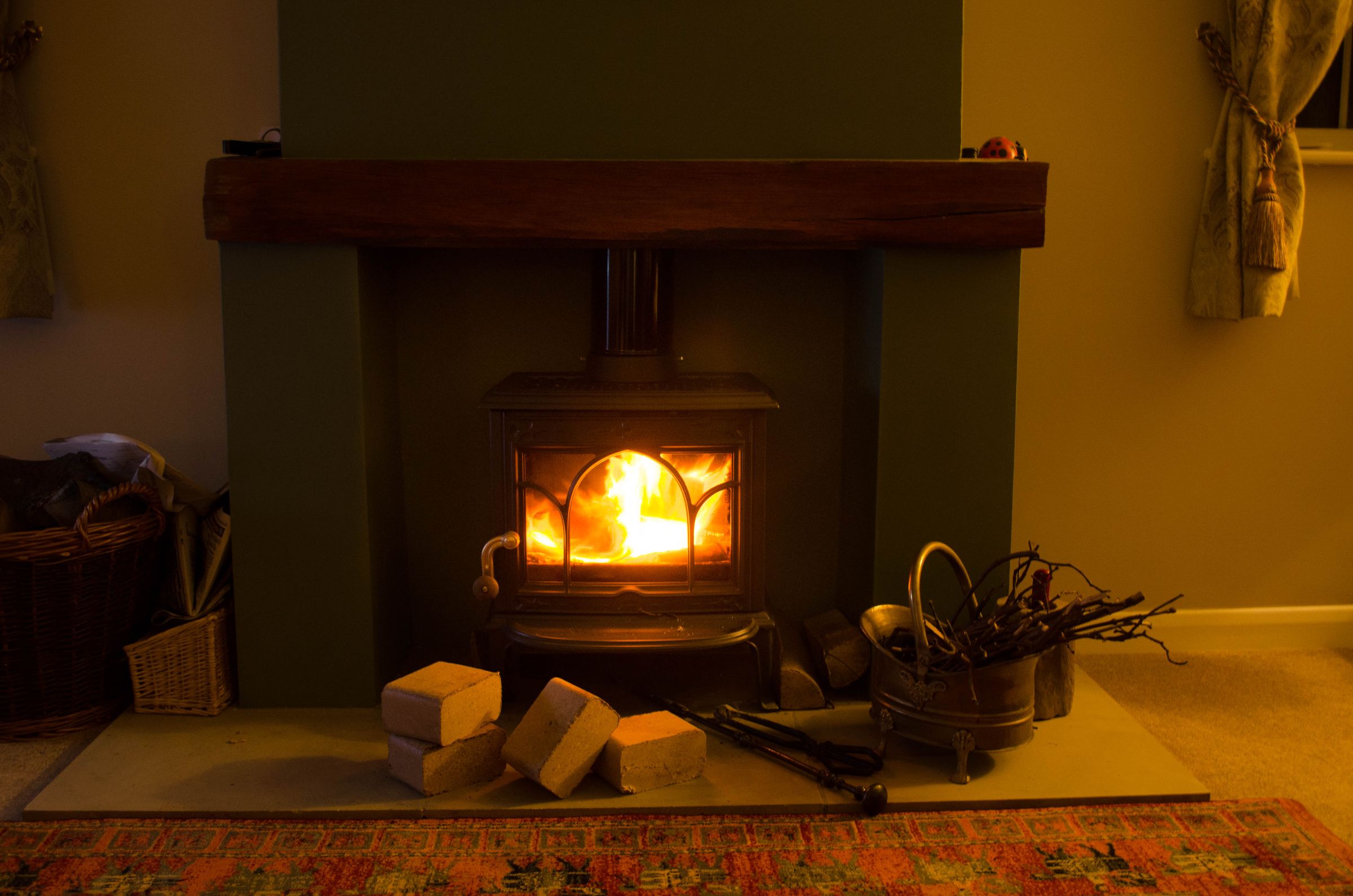 Wood briquettes stacked on the hearth beside a lit stove under an oak beam mantel